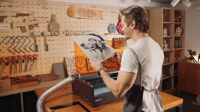 Maker holding up a photo-engraved dog portrait beside an enclosed AtomStack Kraft with an exhaust hose running to a window